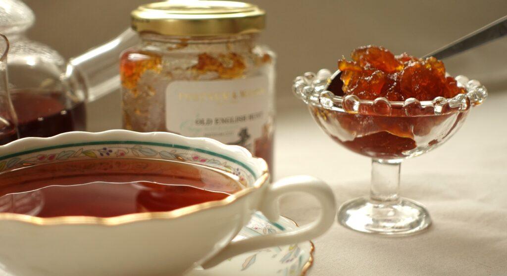 Photo of a glass compotière filled with marmalade, with a jar of Fortnum & Mason marmalade in the background and an elegant cup of tea in the foreground, capturing a refined tea-time setting.