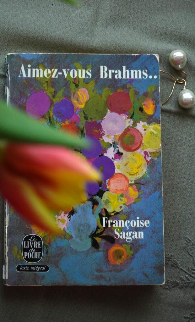 Photo of the novel Aimez-vous Brahms by Françoise Sagan, with a pair of pearl earrings placed beside it and a softly blurred tulip in the foreground adding a touch of color and depth.