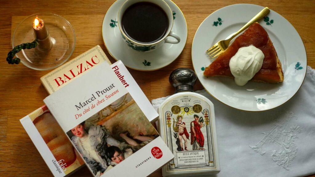 Flat lay of a Proust novel, a bottle of Buly scented body oil, a cup of coffee and a slice of tarte tatin