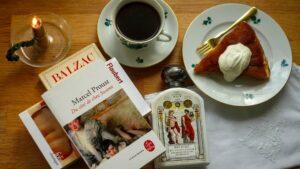 Flat lay of a Proust novel, a bottle of Buly scented body oil, a cup of coffee and a slice of tarte tatin