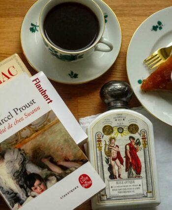 Flat lay of a Proust novel, a bottle of Buly scented body oil, a cup of coffee and a slice of tarte tatin