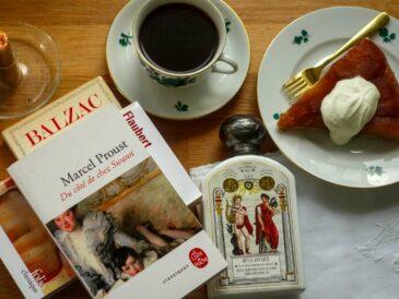 Flat lay of a Proust novel, a bottle of Buly scented body oil, a cup of coffee and a slice of tarte tatin