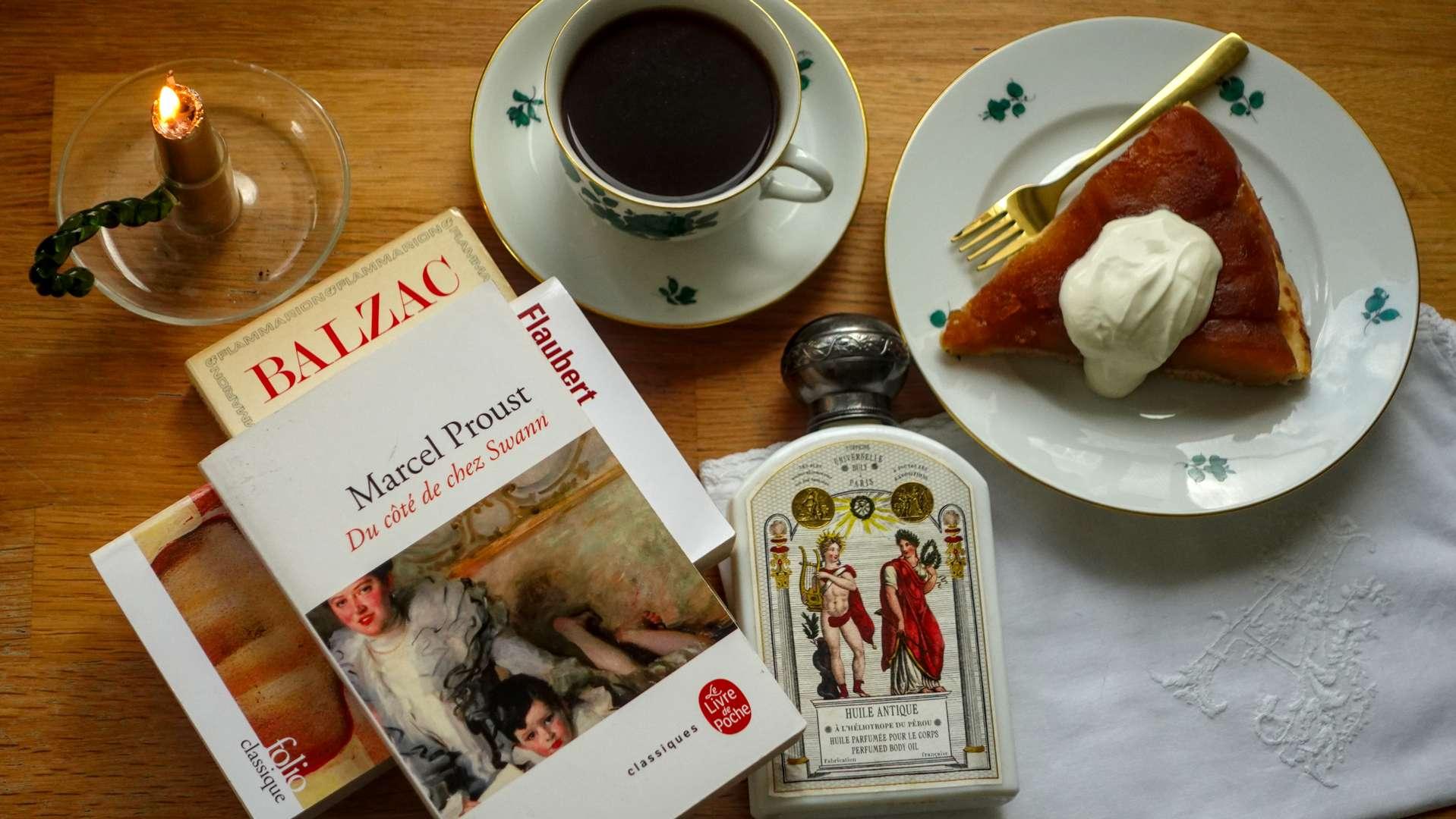 Flat lay of a Proust novel, a bottle of Buly scented body oil, a cup of coffee and a slice of tarte tatin