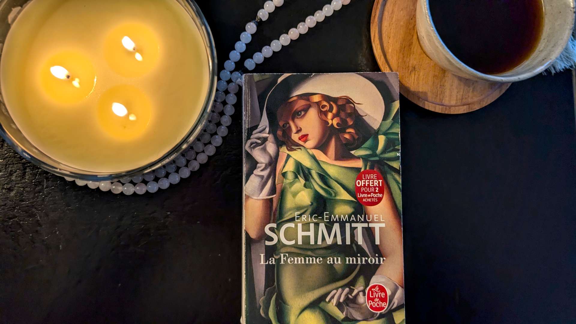 A copy of Eric-Emmanuel Schmitt's novel Three Women in a Mirror (La Femme au miroir) as a quiet reading ritual with coffee and candles.