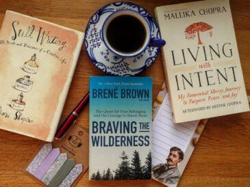 A top-down flat lay of three books: Living with Intent by Mallika Chopra, Braving the Wilderness by Brené Brown, and Still Writing by Dani Shapiro, arranged neatly on a wooden surface.