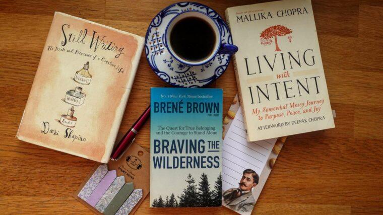 A top-down flat lay of three books: Living with Intent by Mallika Chopra, Braving the Wilderness by Brené Brown, and Still Writing by Dani Shapiro, arranged neatly on a wooden surface.