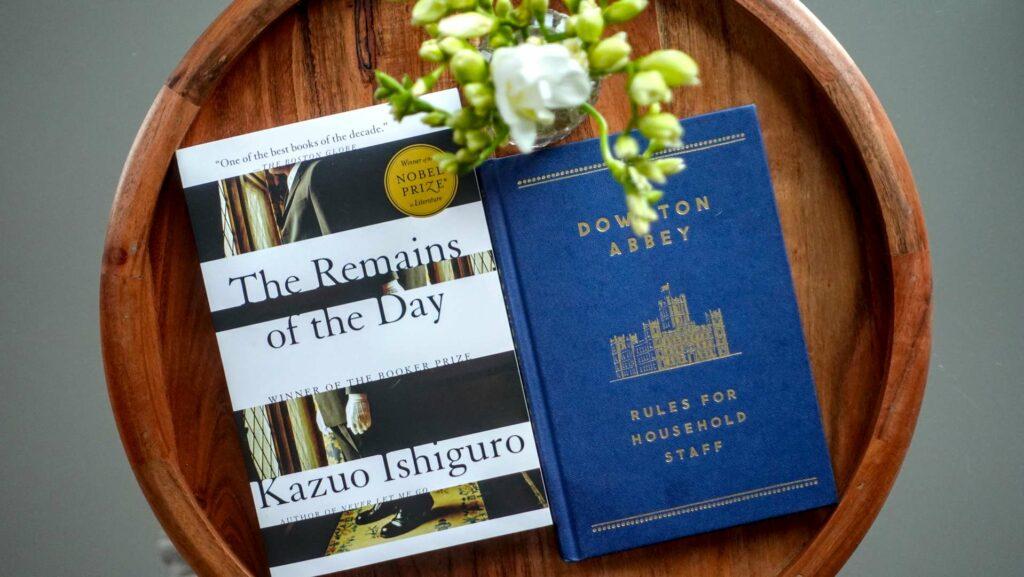 A literary flatlay of The Downton Abbey Rules for Household Staff and Kazuo Ishiguro’s The Remains of the Day next to a small bouquet of white freesia, exploring themes of dignity and service.