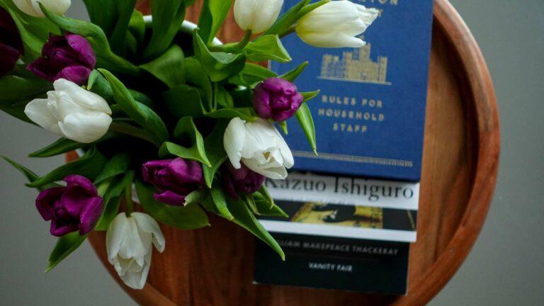 A top-down flatlay on a wooden tray featuring books about modern etiquette and literature, including The Downton Abbey Rules for Household Staff, The Remains of the Day, and Vanity Fair, accented with fresh tulips.