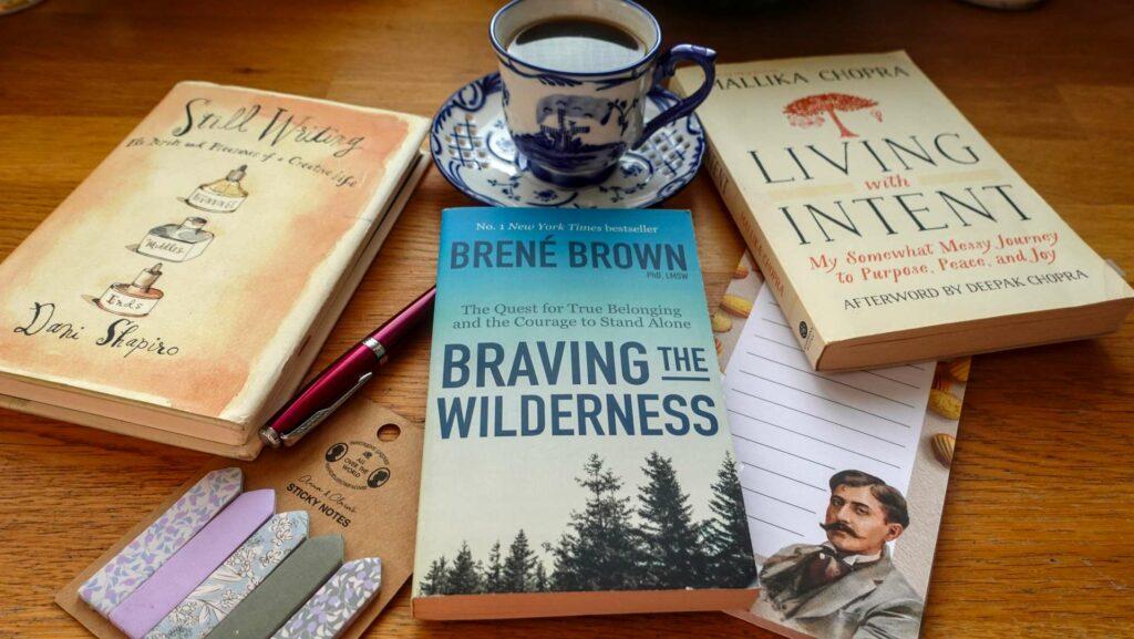 A top-down flat lay of three books: Living with Intent by Mallika Chopra, Braving the Wilderness by Brené Brown, and Still Writing by Dani Shapiro, arranged neatly on a wooden surface.