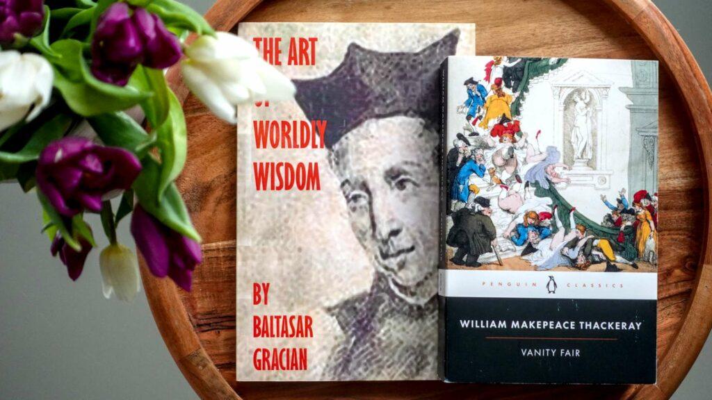 A flatlay of Vanity Fair by Thackeray and The Art of Worldly Wisdom by Baltasar Gracián with pink tulips, representing the intersection of modern etiquette and literature.