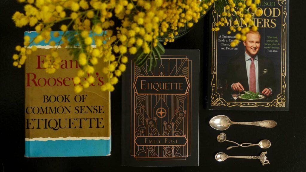A literary collection of etiquette books by Emily Post, Eleanor Roosevelt, and William Hanson arranged with a yellow branch of mimosa blooms on a soft background.