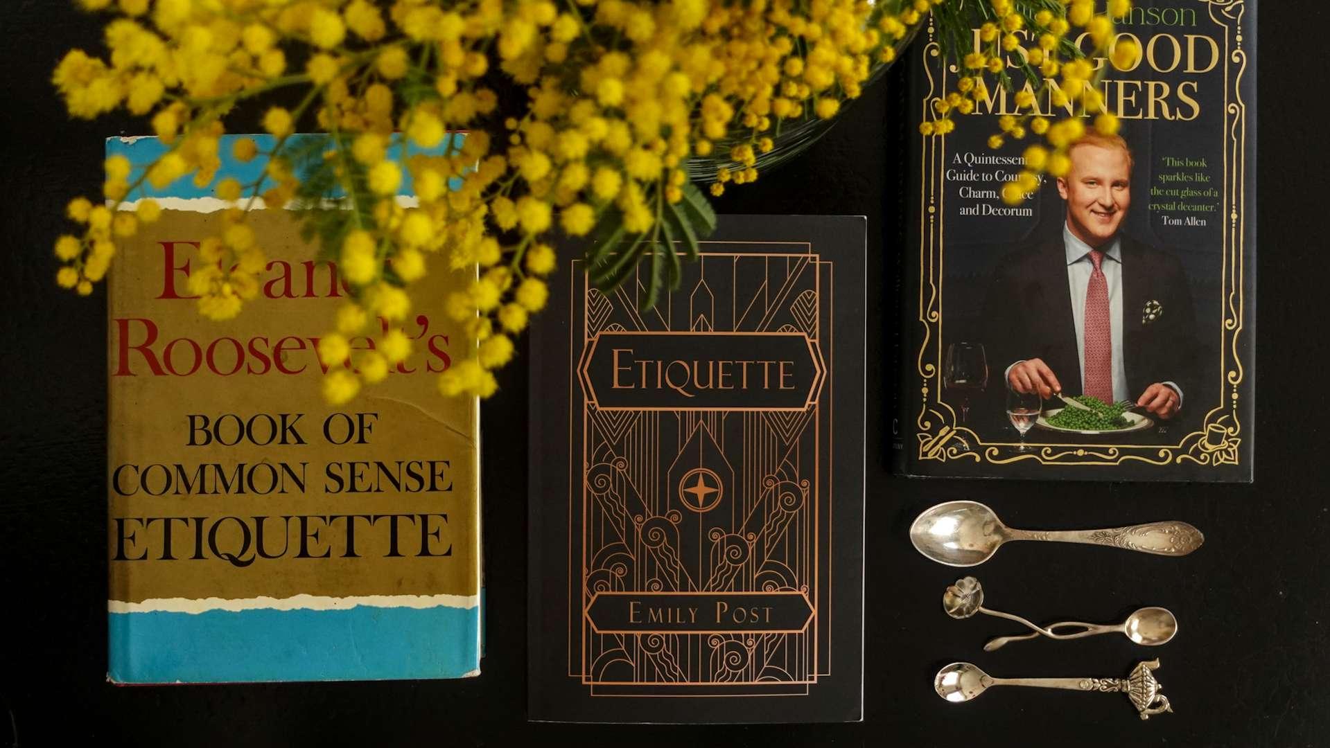 A literary collection of etiquette books by Emily Post, Eleanor Roosevelt, and William Hanson arranged with a yellow branch of mimosa blooms on a soft background.