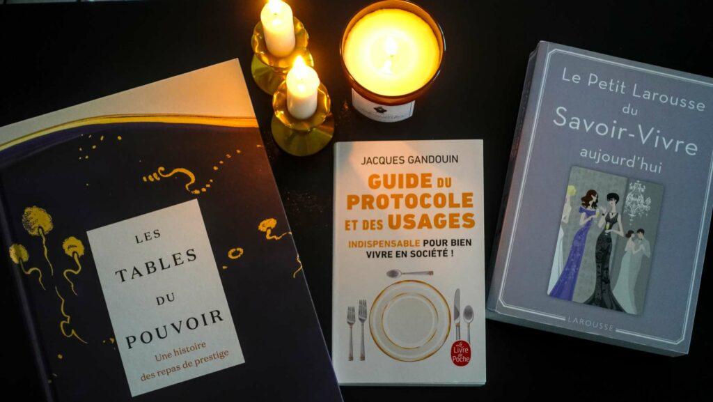 A cozy flatlay of three French etiquette books surrounded by lit candles on a lacquered wooden table.