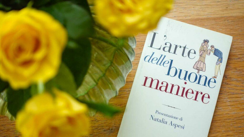 The Italian book L'arte delle buone maniere by Il Mulino on a wooden surface with a floral arrangement.