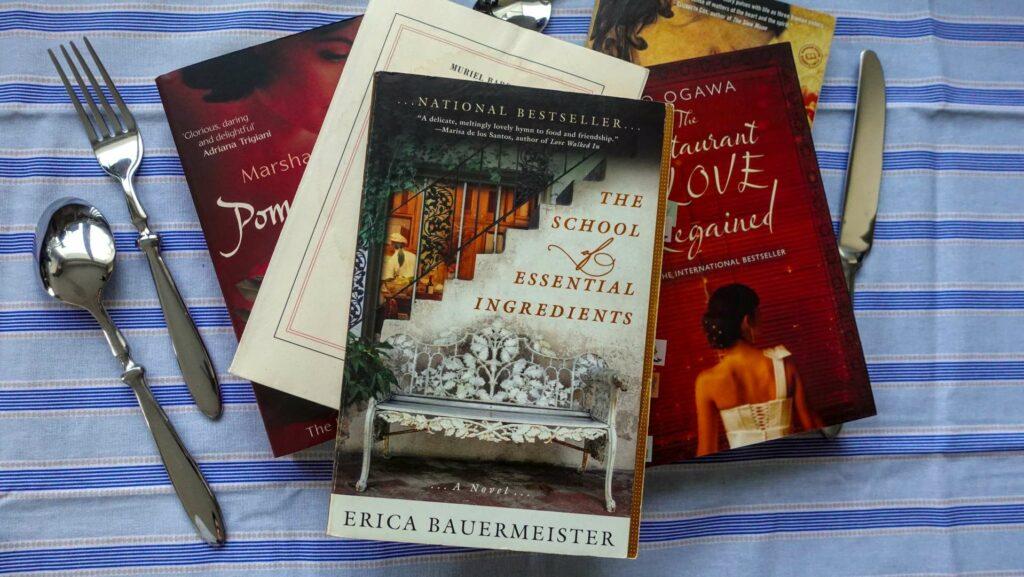 A cozy stack of food-themed novels sitting on a rustic kitchen counter next to some cutlery on a striped tablecloth