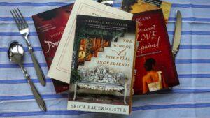 A cozy stack of food-themed novels sitting on a rustic kitchen counter next to some cutlery on a striped tablecloth