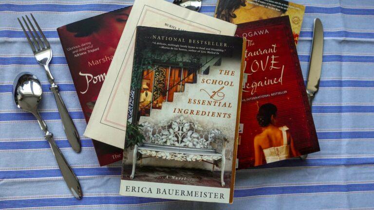 A cozy stack of food-themed novels sitting on a rustic kitchen counter next to some cutlery on a striped tablecloth