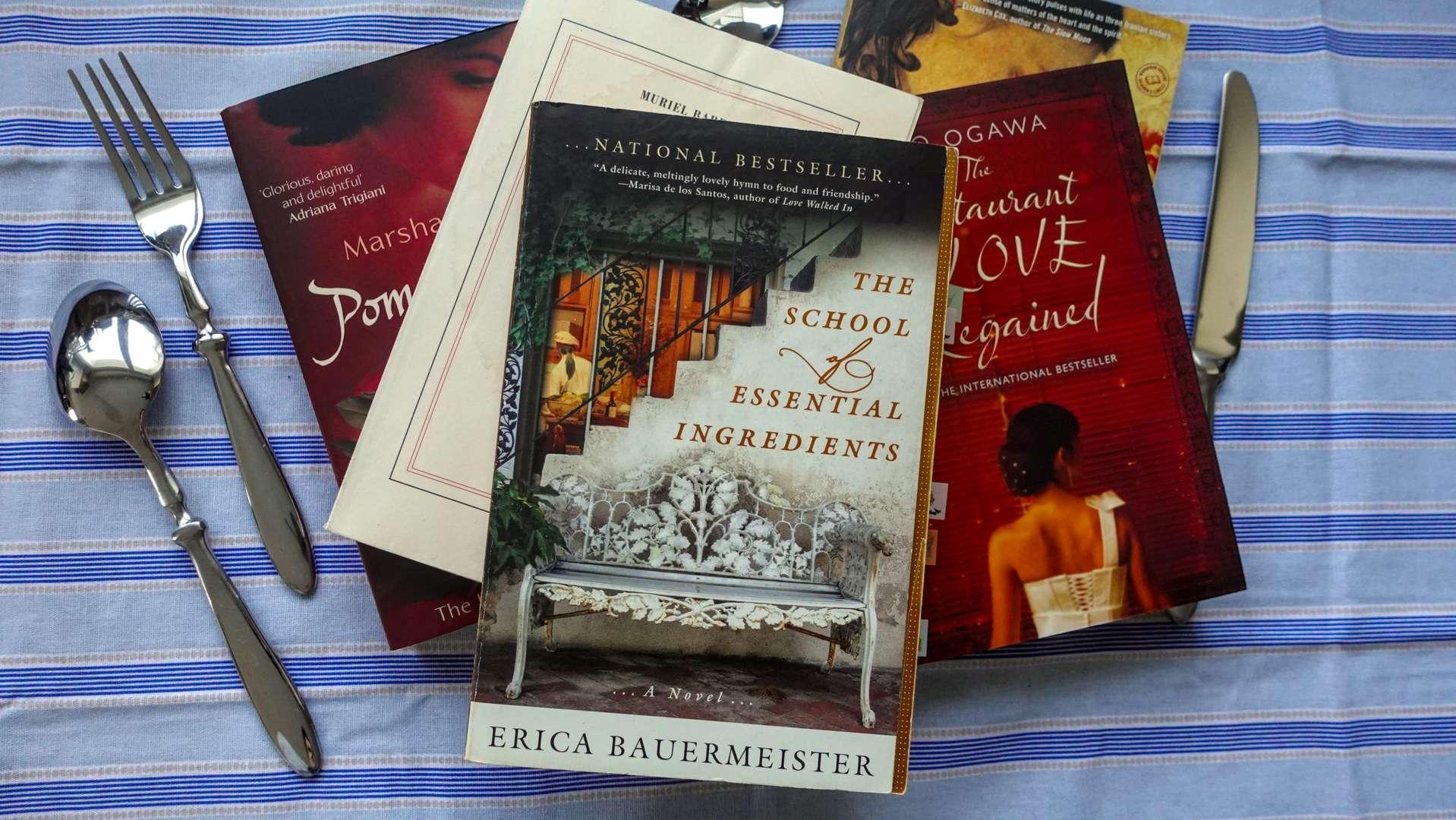 A cozy stack of food-themed novels sitting on a rustic kitchen counter next to some cutlery on a striped tablecloth