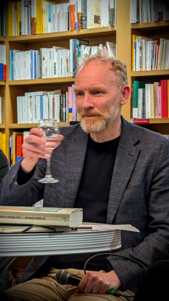Author Jón Kalman Stefánsson speaking at a literary event in a bookshop, holding a glass of water with bookshelves in the background.
