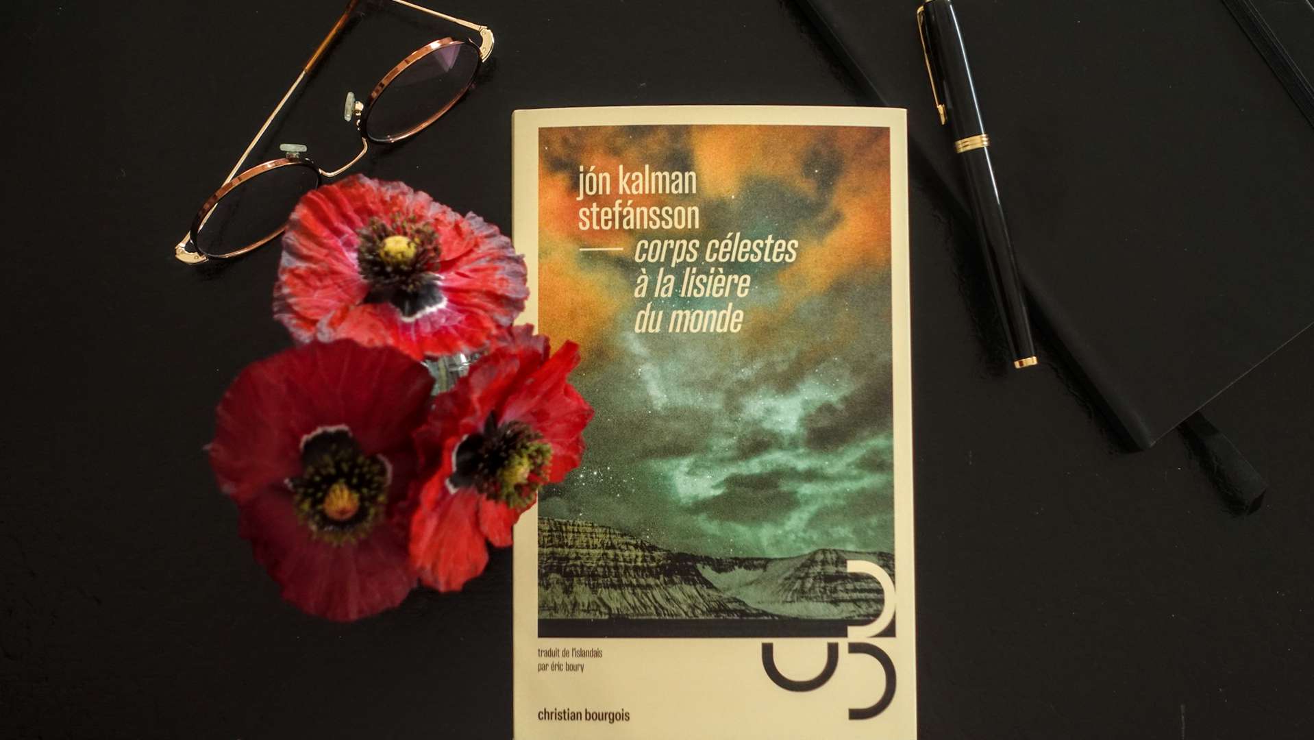 Front cover of Jón Kalman Stefánsson’s novel "Corps célestes à la lisière du monde" on a black table next to a pair of glasses and red poppy flowers.