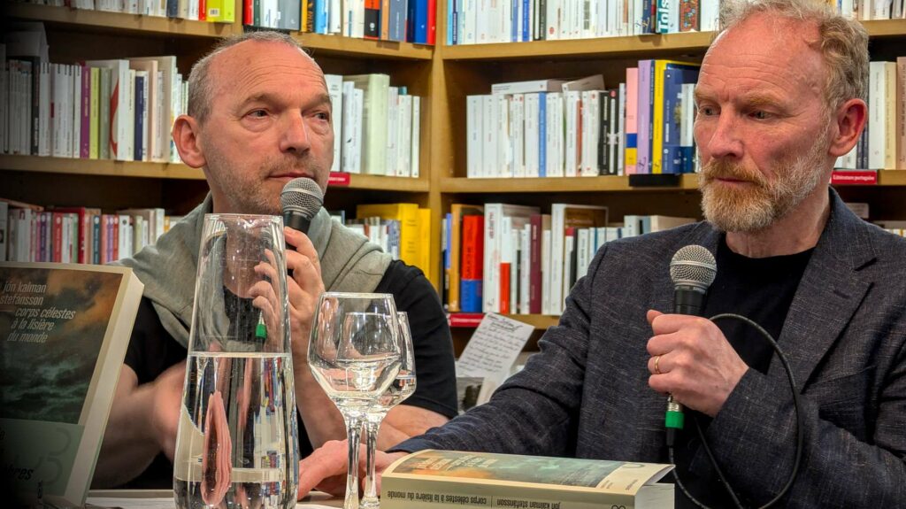 Jón Kalman Stefánsson and an interpreter holding microphones during a French book launch, surrounded by books and a carafe of water.