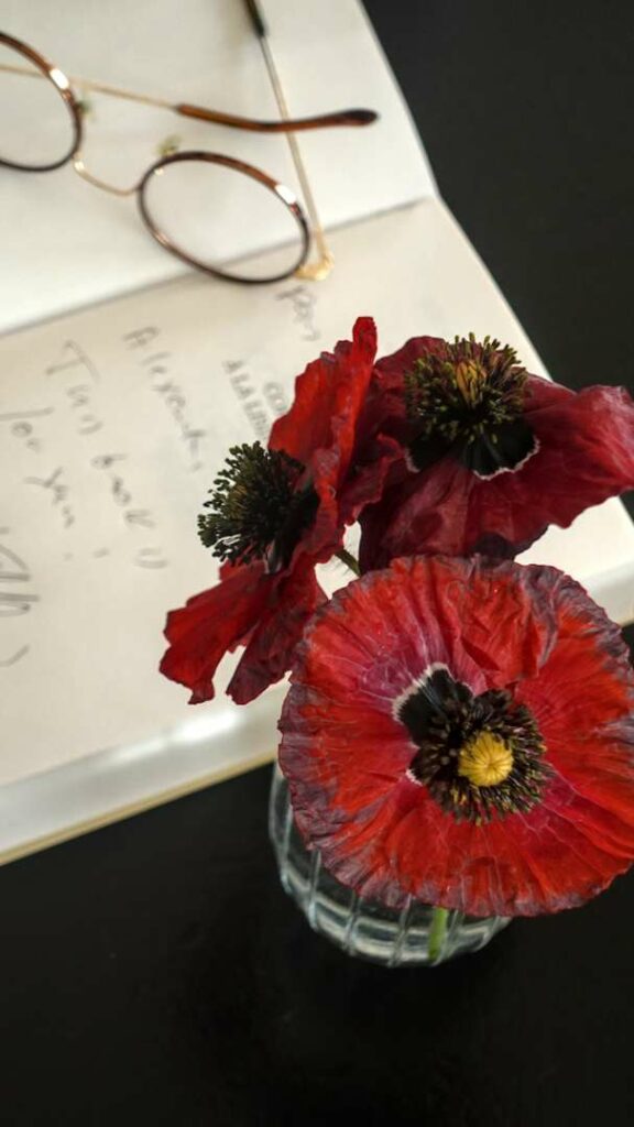 Close-up of three vibrant red poppies in a small glass vase, positioned in front of a signed book and glasses on a dark surface.