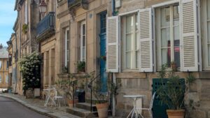 A quiet cobbled street in Autun lined with stone facades, white shutters, potted plants and wrought iron café chairs