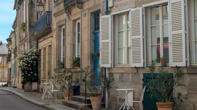 A quiet cobbled street in Autun lined with stone facades, white shutters, potted plants and wrought iron café chairs