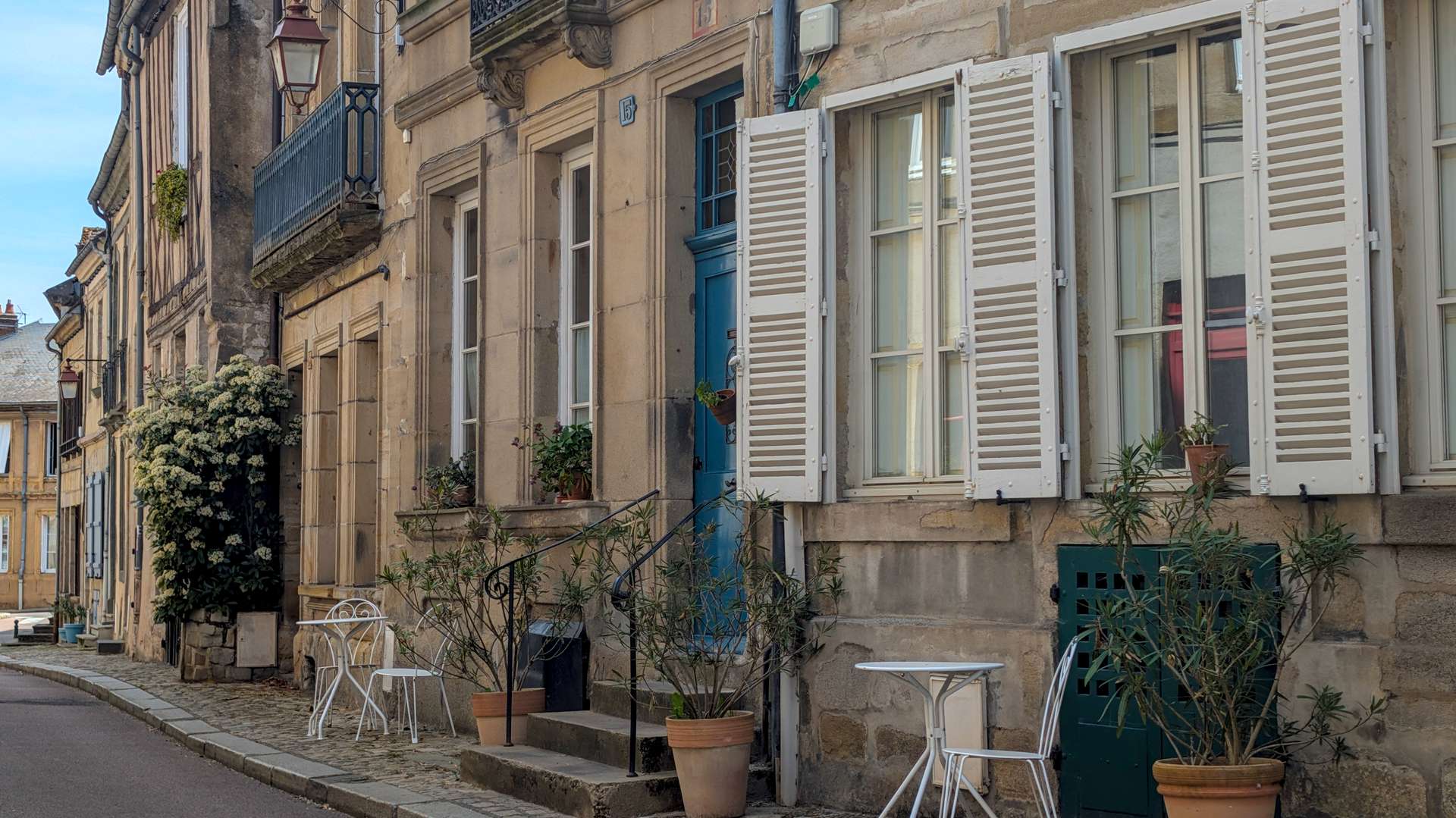 A quiet cobbled street in Autun lined with stone facades, white shutters, potted plants and wrought iron café chairs