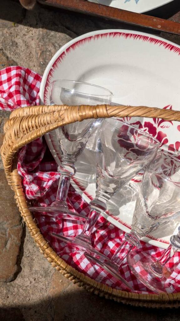 A wicker basket lined with red gingham cloth holding crystal wine glasses and a white plate edged in red, found in a brocante in Autun