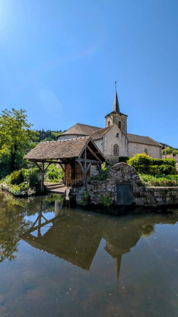 A small Burgundian church and its wooden lavoir perfectly reflected in still water under a clear blue sky