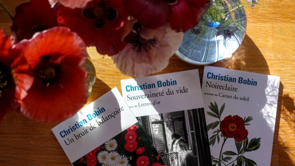 Three books by Christian Bobin fanned out on a wooden table with red flowers in a glass vase